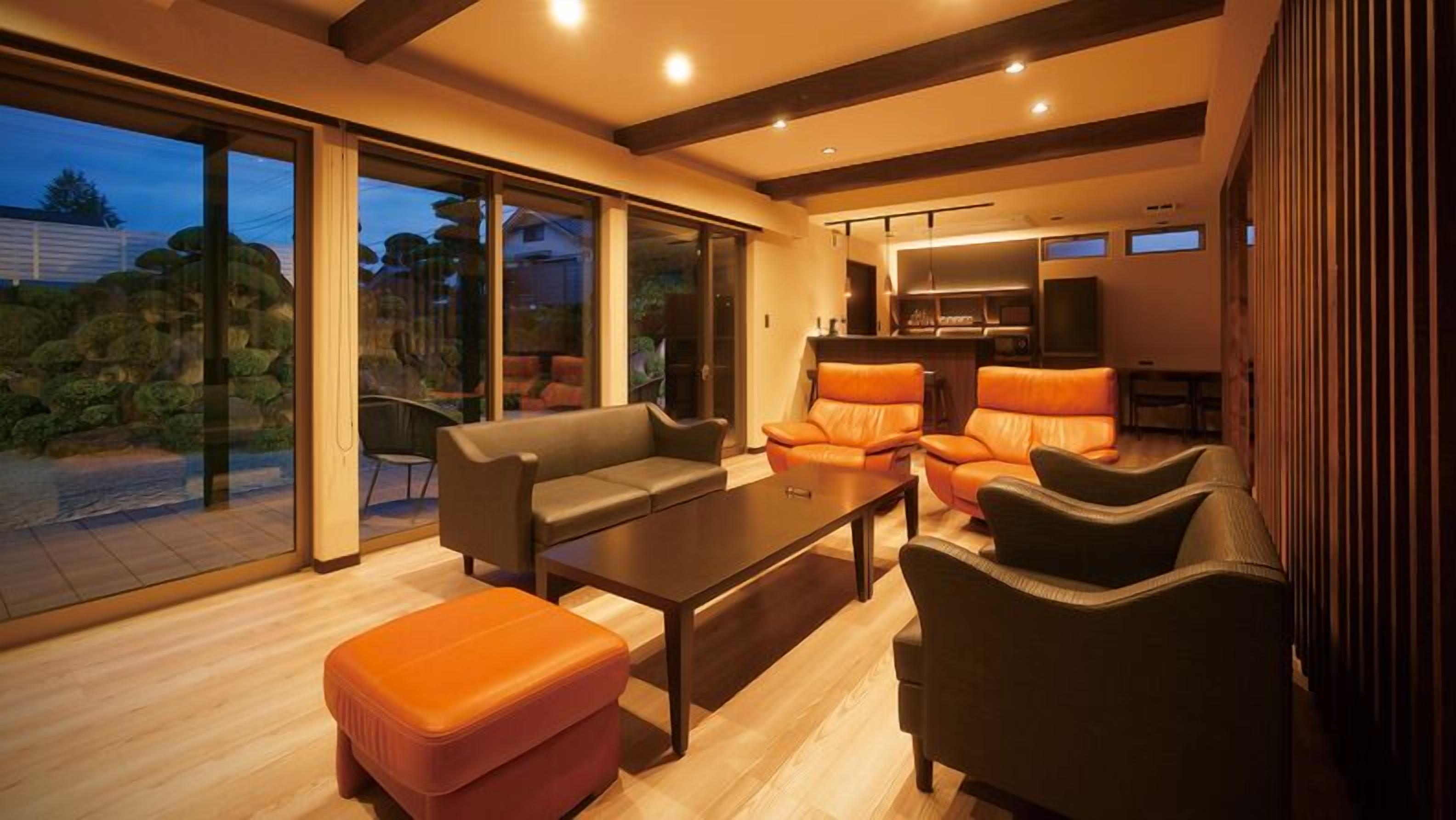 A modern living room with a sofa, ottoman, and chairs, a dining area with a table, and a kitchen with a TV.