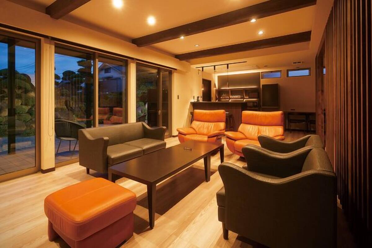 Un salon moderne comprenant un canapé, un pouf et des fauteuils, un coin repas avec une table, et une cuisine avec un téléviseur.