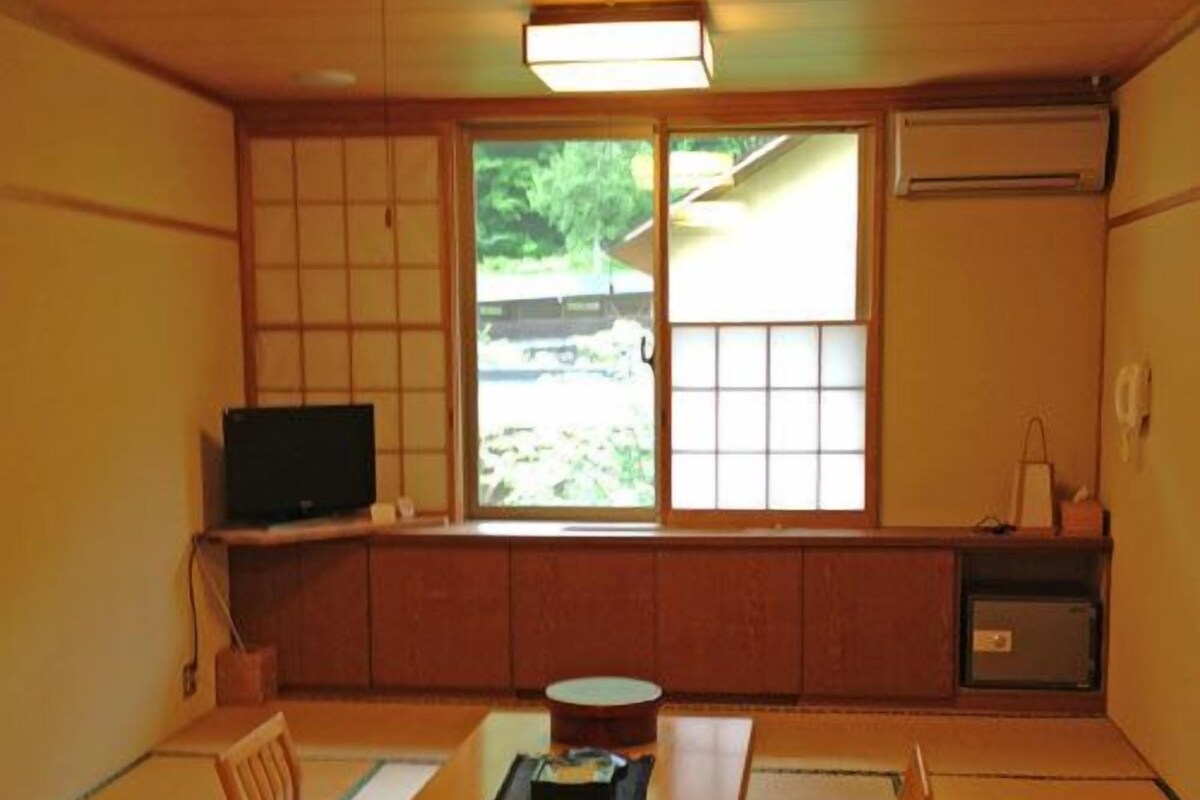 A traditional Japanese room with tatami flooring, a low table with cushions, a television, and sliding doors with a view of greenery.