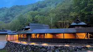 A traditional wooden building with a stone wall in front, set against a forested hillside. - Azuki Onsen Kaboku no Yado (Minamiaizu)