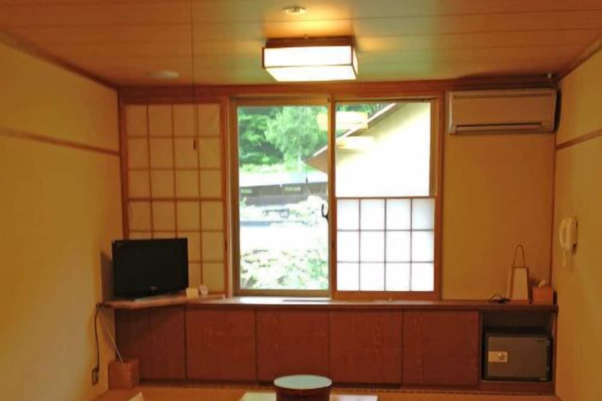 A traditional Japanese room with tatami flooring, a low table with cushions, a television, and sliding doors with a view of greenery.