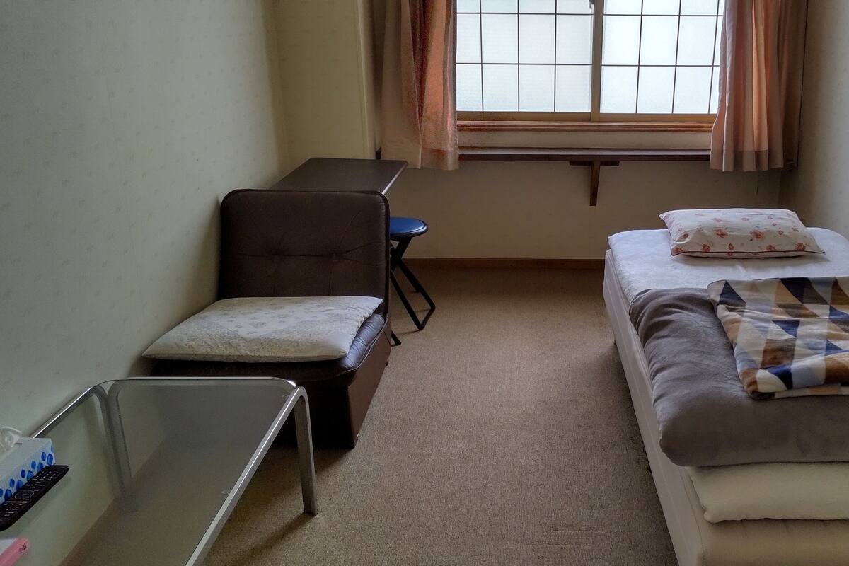 A small, sparsely furnished room with a bed, a chair, a desk, and a window with curtains.