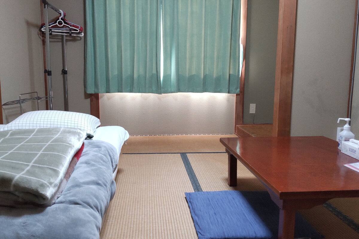 A hotel room with a bed, a wooden table, a chair, a mirror, and a window with curtains.