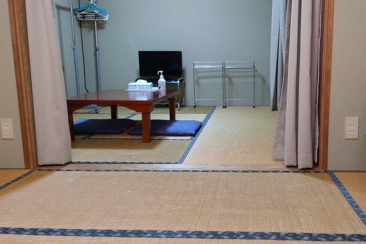 A room with a bed, a table with a chair, a television, and a small table with a bottle and tissues.