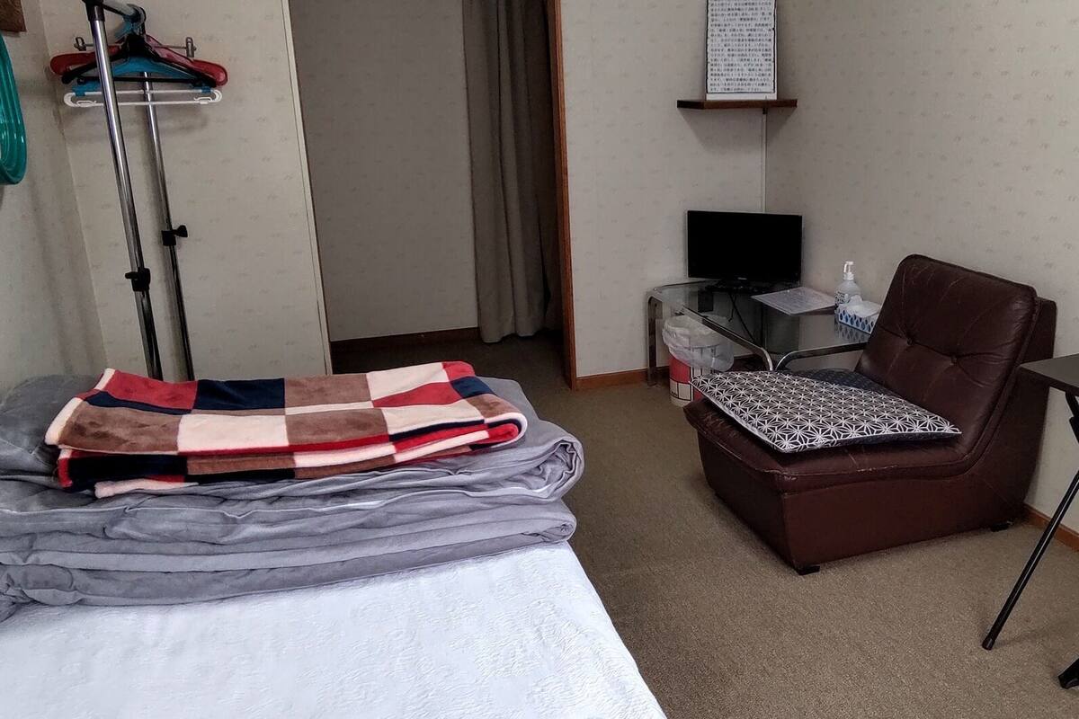 A hotel room with a bed, a chair, a small table, a TV, and a wall-mounted shelf.