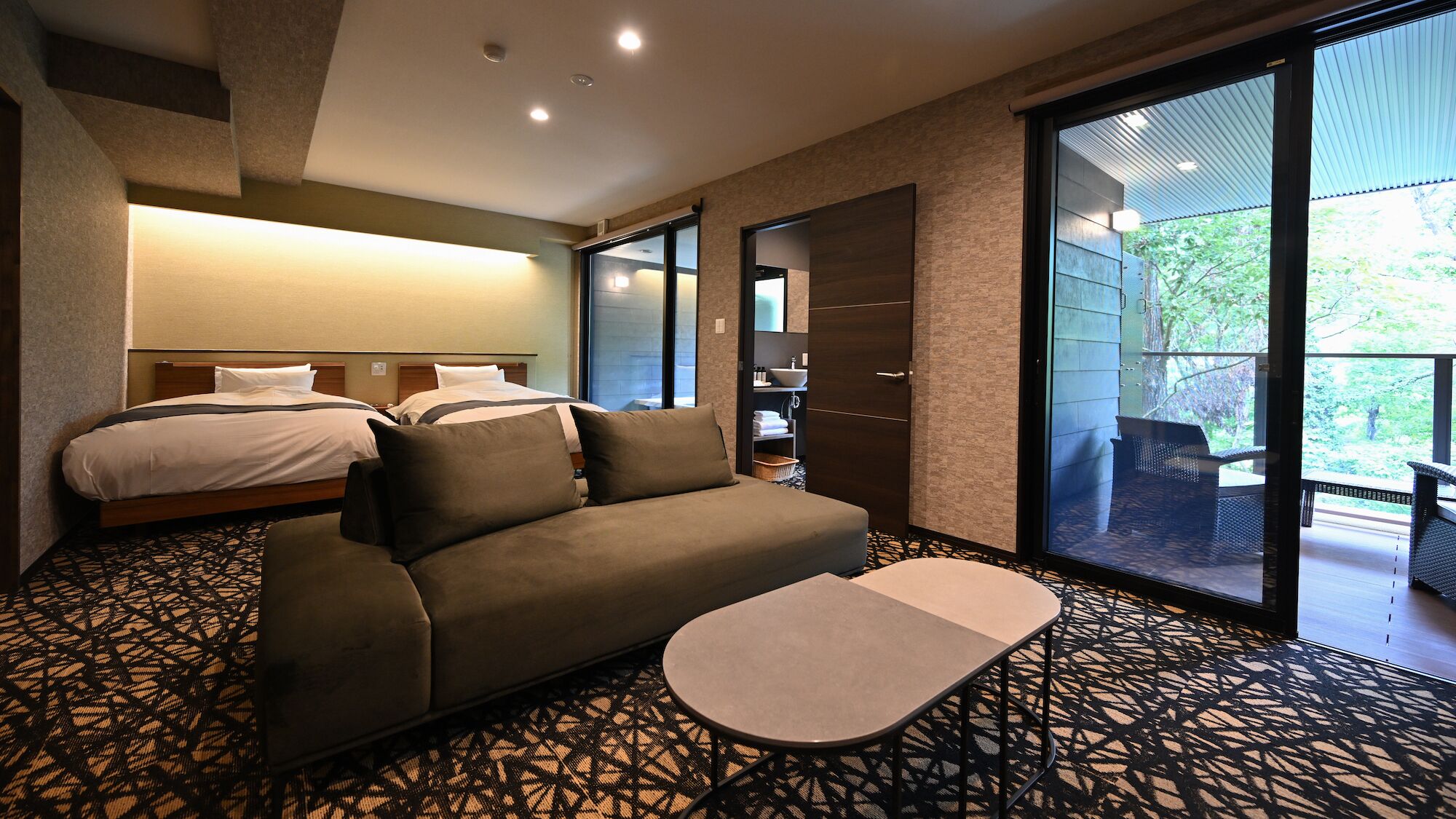 Ein Hotelzimmer mit Bett, Sofa und einem kleinen Tisch. Eine Schiebetür aus Glas führt zu einem Außenbereich.