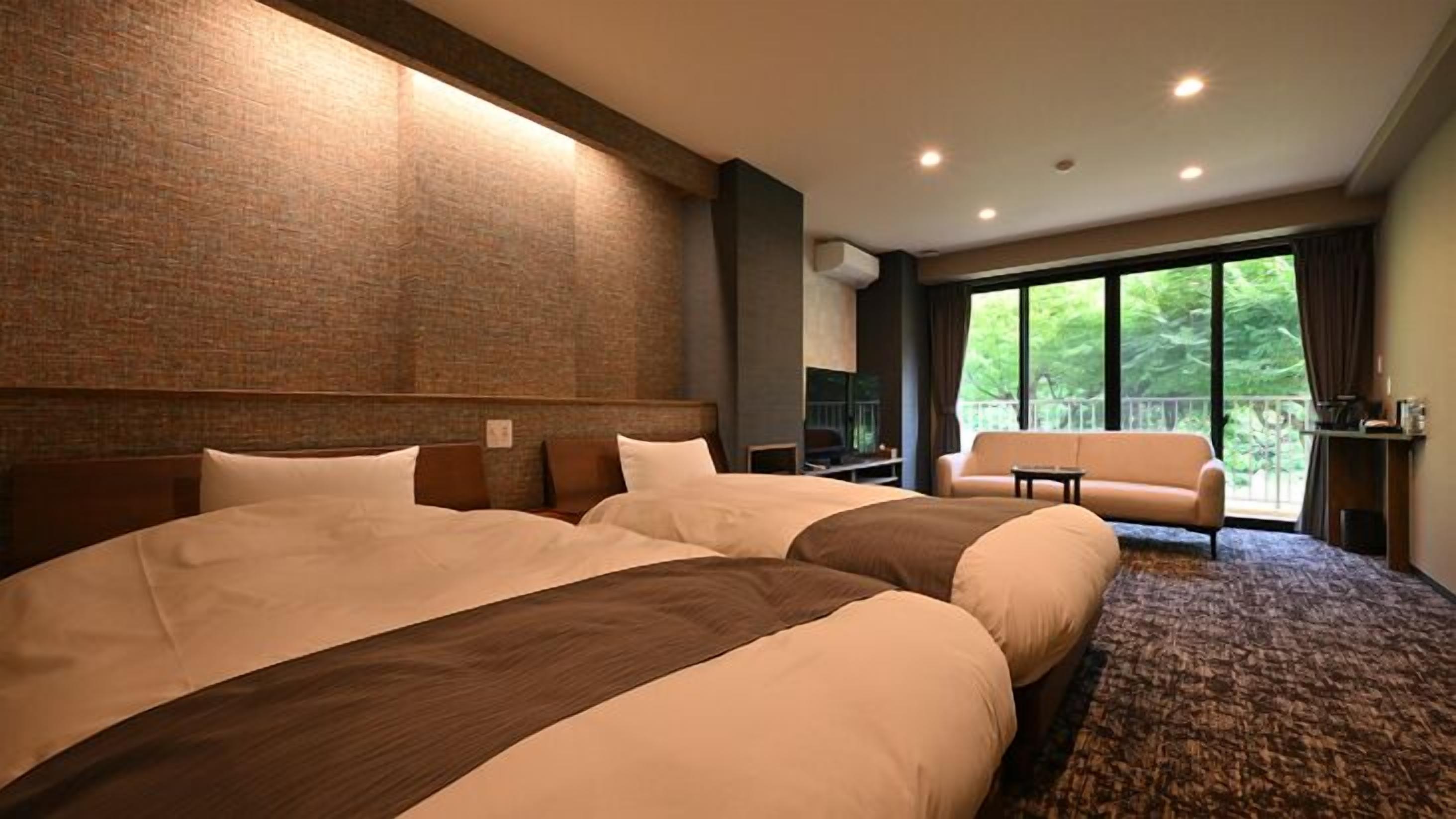 A hotel room with two large beds, a sofa, a balcony with a view of greenery, and a brick wall.