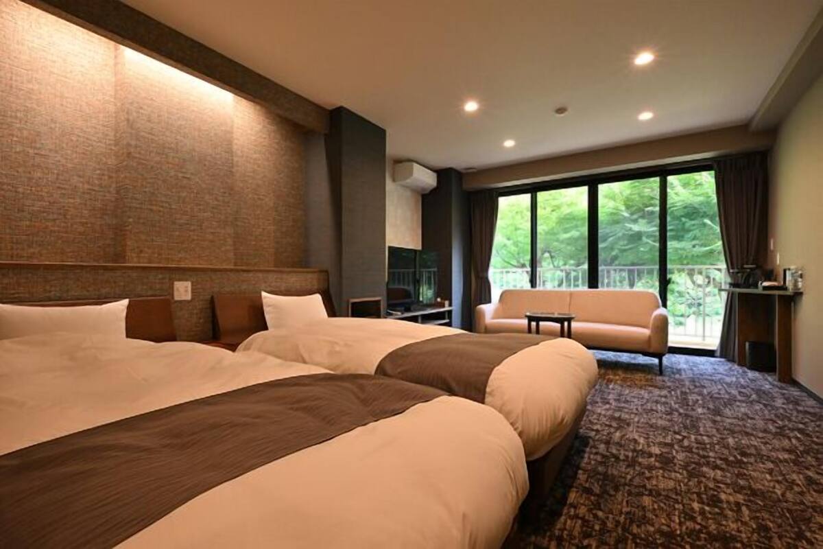 Ein Hotelzimmer mit zwei grossen Betten, einem Sofa, einem Balkon mit Blick auf Grünflächen und einer Ziegelwand.