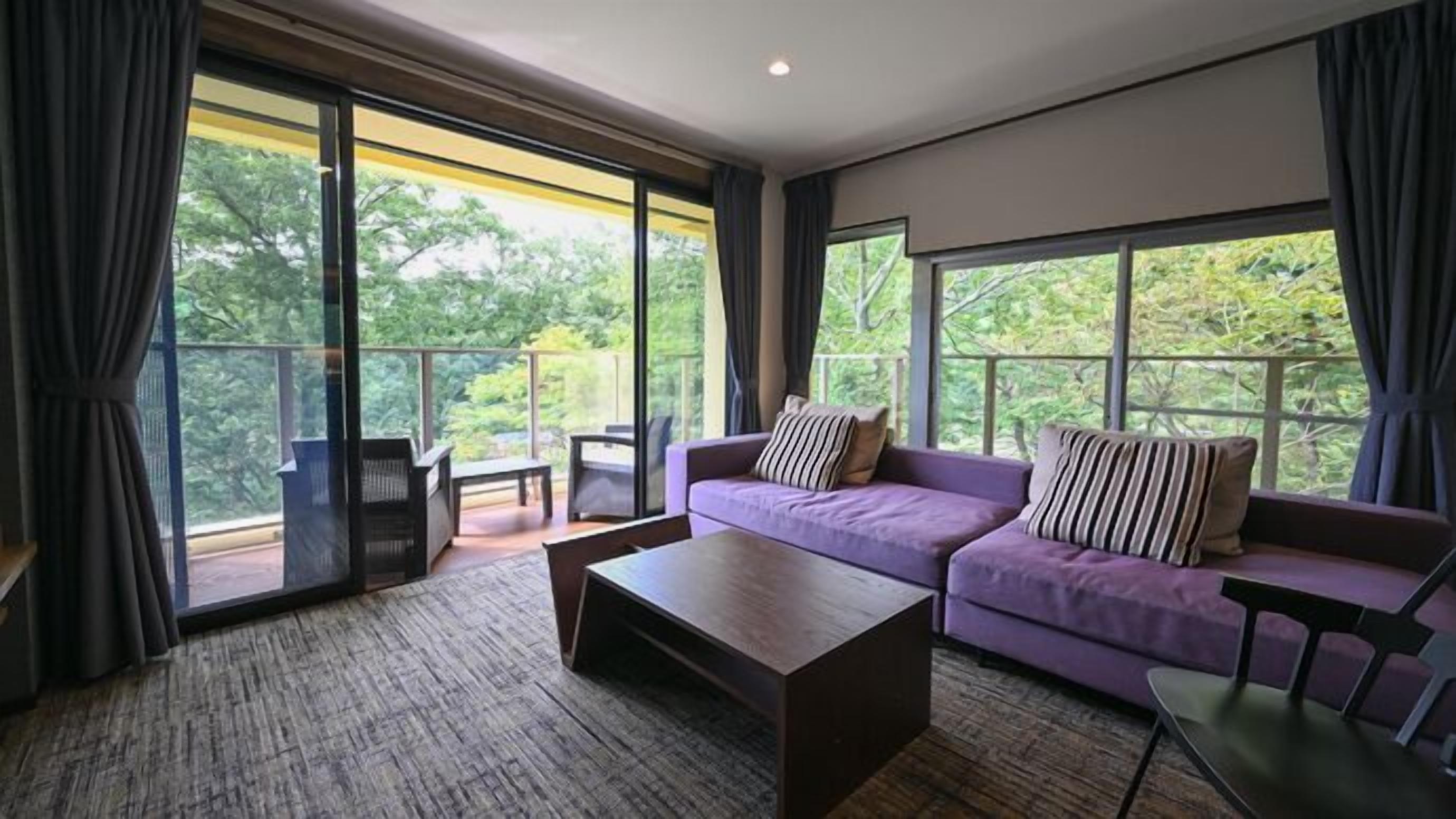 A modern living room with a sofa, coffee table, and a view of greenery through large windows.