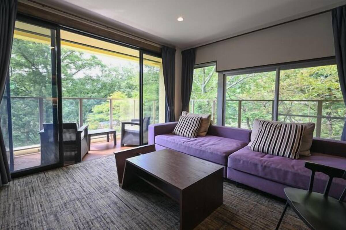 Ein modernes Wohnzimmer mit einer Couch, einem Couchtisch und Blick auf Grün hinter großen Fenstern.