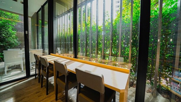 A modern dining area with a long table and chairs, large windows offering a view of greenery and buildings. - Quintessa Hotel Tokyo Haneda Comic & Books (Tokyo)