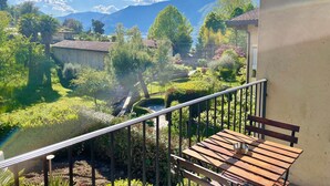 A balcony with a wooden bench and a metal railing overlooking a lush garden with a mountain in the background.