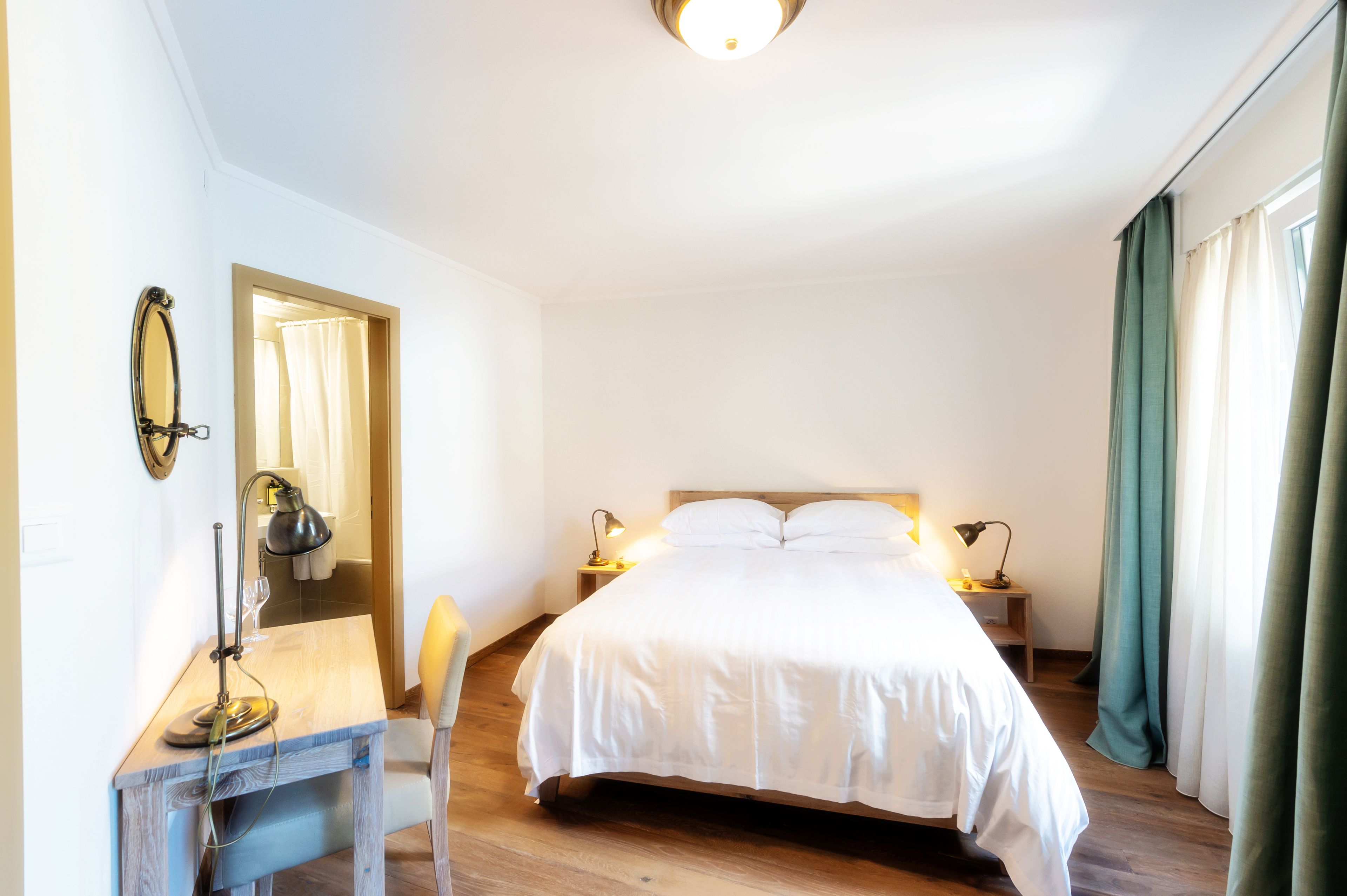 Une chambre à coucher avec un lit, des lampes de chevet, un bureau avec une lampe, un miroir et une fenêtre avec des rideaux.
