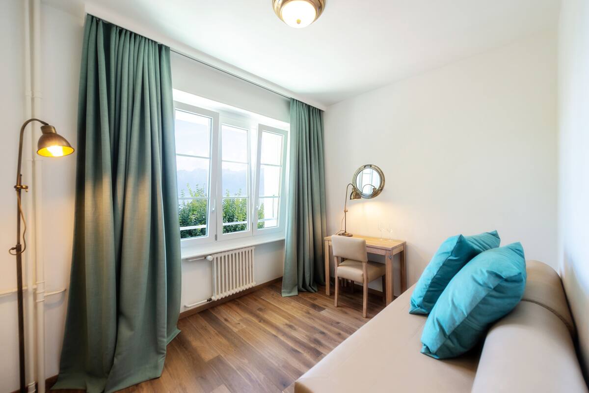 Une pièce avec un plancher en bois, un lit avec des oreillers bleus, un bureau avec une chaise, une fenêtre avec des rideaux et une lampe.