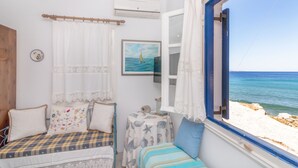 A bedroom with a bed, a small table, a window with curtains, a painting, and a view of the sea. - Luxury Wave  Maisonette Ormos Isternion Tinos (Tinos)