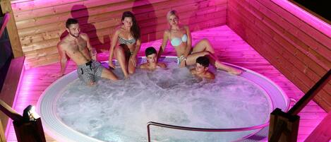A hot tub with four people sitting inside, surrounded by wooden walls and pink lighting.