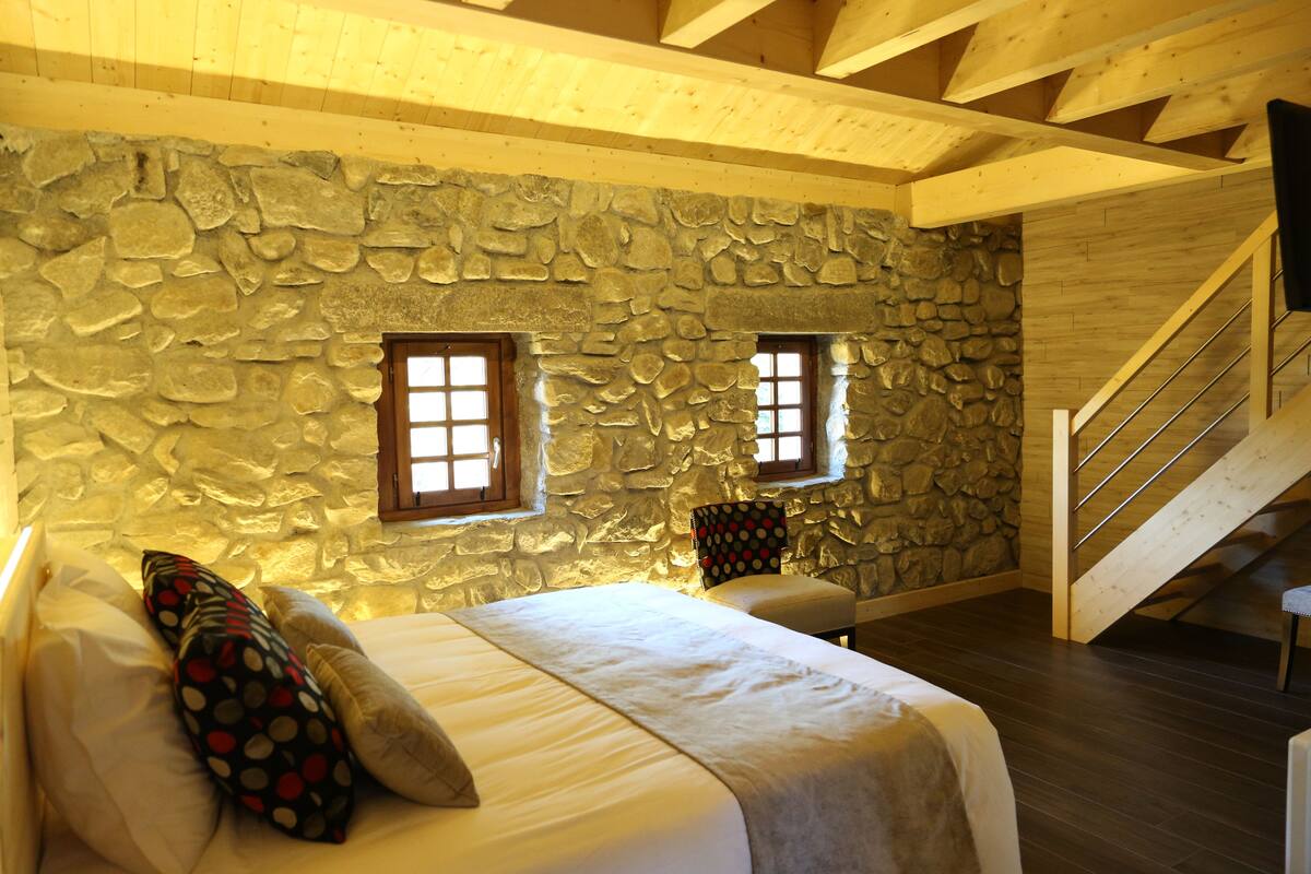 A bedroom with a stone wall, wooden beams, a bed with pillows, a chair, and a staircase.