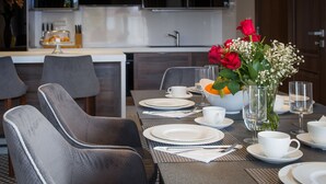 Un coin repas avec une table mise pour quatre personnes, comprenant des assiettes, des tasses et des couverts. On peut y voir un vase avec des fleurs et un saladier rempli de fruits.