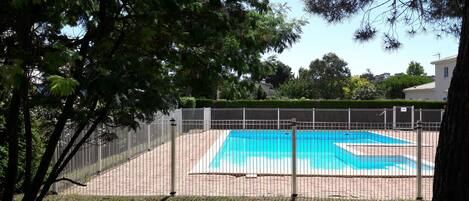 A fenced swimming pool area with a clear blue pool, surrounded by a brick pathway and greenery.