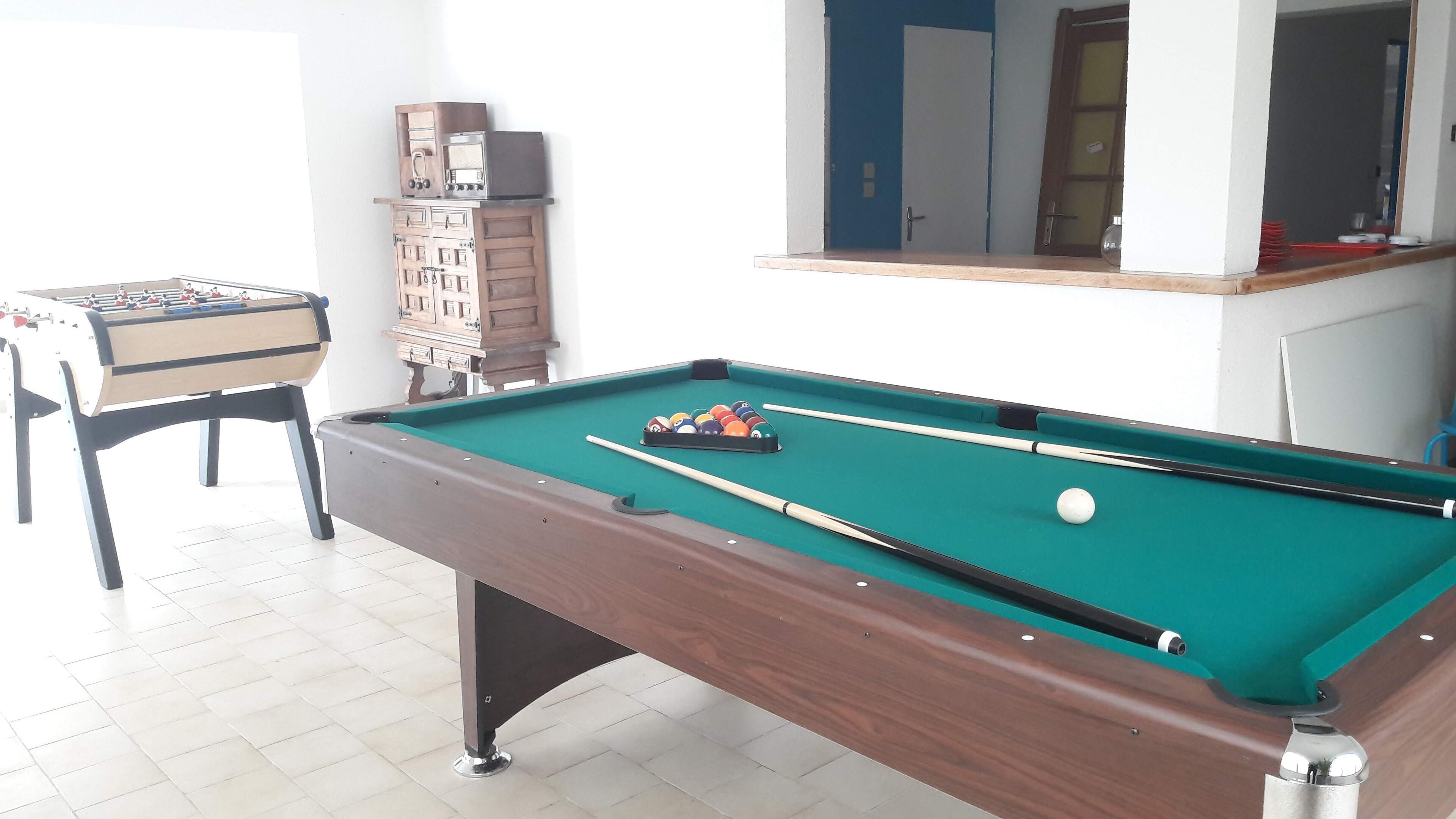Ein Billardtisch mit grüner Spielbahn, ein Schusszepter und ein Satz Billardkugeln. Links daneben steht ein Kicker und dahinter ein Pool Tisch eine Holzkabine mit Schubladen.