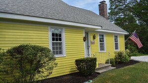 Une maison jaune avec une porte blanche, des fenêtres et une cheminée.