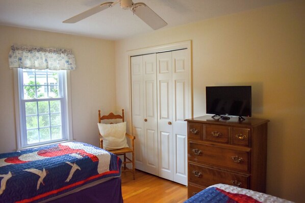 A bedroom with two beds, a wooden dresser, a television, a chair, and a white door. - Charming, well appointed home has hardwood floors and ceiling fans throughout (West Harwich)