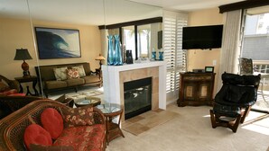 A living room with a fireplace, a sofa, a chair, a television, and a large window with blinds. - Great corner condo! Surrounding Ocean Views - D2 North Coast Village (Oceanside)