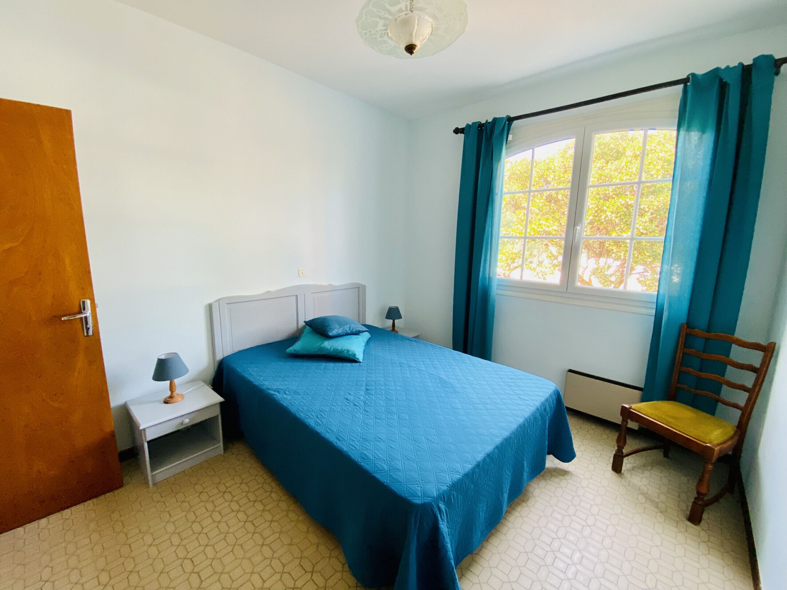 Un dormitorio con cama, mesita de noche, una silla, una ventana con cortinas y una puerta.