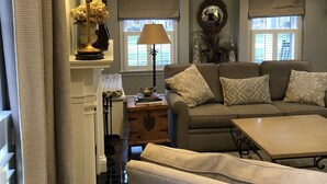A cozy living room with a sofa, a coffee table, and a lamp. There are decorative items on a shelf and a window with shutters. - Kentucky at its best! Historical meets modern amenities! (Louisville)