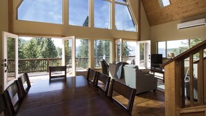 A spacious living area with a large wooden table, several chairs, a sofa, and a TV. The room has large windows offering views of a forested area. - Carlin Bay Retreat on Lake Coeur d'Alene  (Harrison)