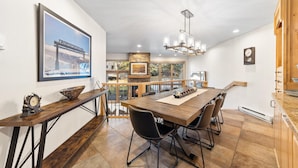 A spacious dining area with a wooden table, black chairs, and a chandelier. A stone fireplace and a large window with a view are visible in the background. - Creekside, multi level condo with a warm cabin feel, open living space & access to all Grand Hyatt Amenities (Vail)