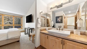 A modern bathroom with a large mirror, a sink, and a toilet. There is a window with a view of trees and a bed with a chair in the adjacent room. - Creekside, multi level condo with a warm cabin feel, open living space & access to all Grand Hyatt Amenities (Vail)