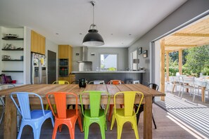 Cozinha moderna com mesa de jantar de madeira e cadeiras coloridas, forno embutido e uma grande janela com vista para um jardim.