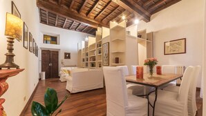 Une pièce spacieuse avec un plafond en bois, des murs blancs et un coin repas comprenant une table et des chaises. On aperçoit des étagères et une porte au fond.