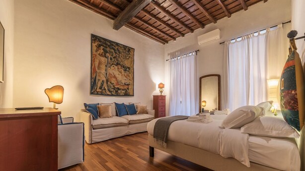 A hotel room with a wooden ceiling, a large painting on the wall, a bed with white linens, a sofa with cushions, and a desk with a lamp. - Unique opportunity to live in a Renaissance palace (Roma)