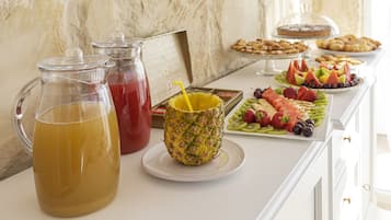 Un bufé de desayuno con diversas frutas, una jarra de jugo de naranja y un vaso de ponche rojo.