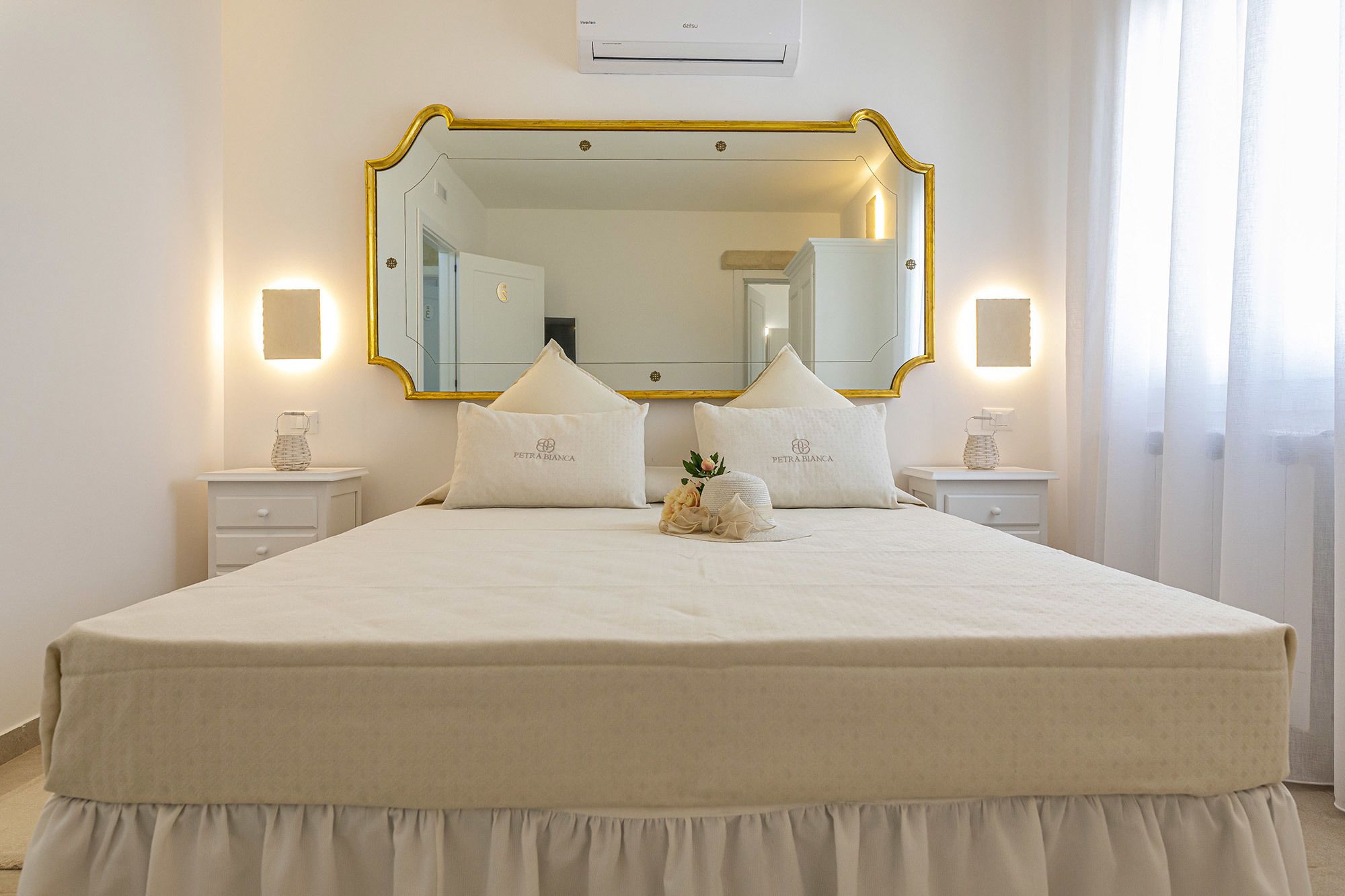 A neatly made bed with a white duvet and pillows, a large mirror with a gold frame, and bedside lamps on white nightstands.