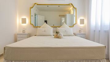 A neatly made bed with a white duvet and pillows, a large mirror with a gold frame, and bedside lamps on white nightstands.