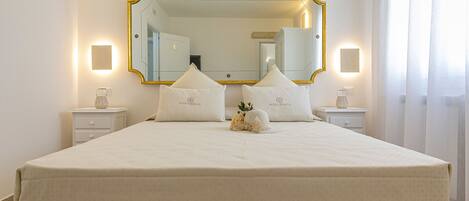 A neatly made bed with a white duvet and pillows, a large mirror with a gold frame, and bedside lamps on white nightstands.
