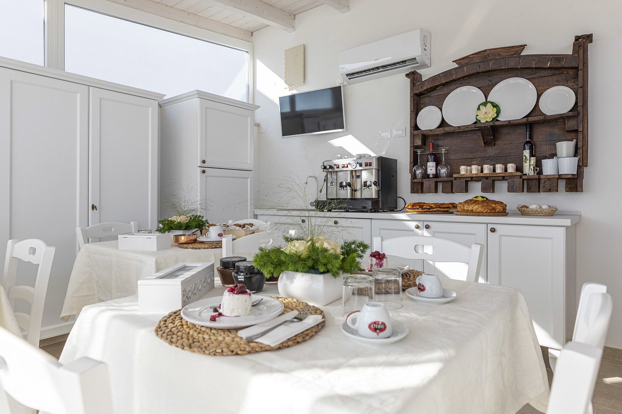 A bright, well-lit kitchen with white cabinetry, a coffee machine, and a rustic wooden shelf with plates and jars.