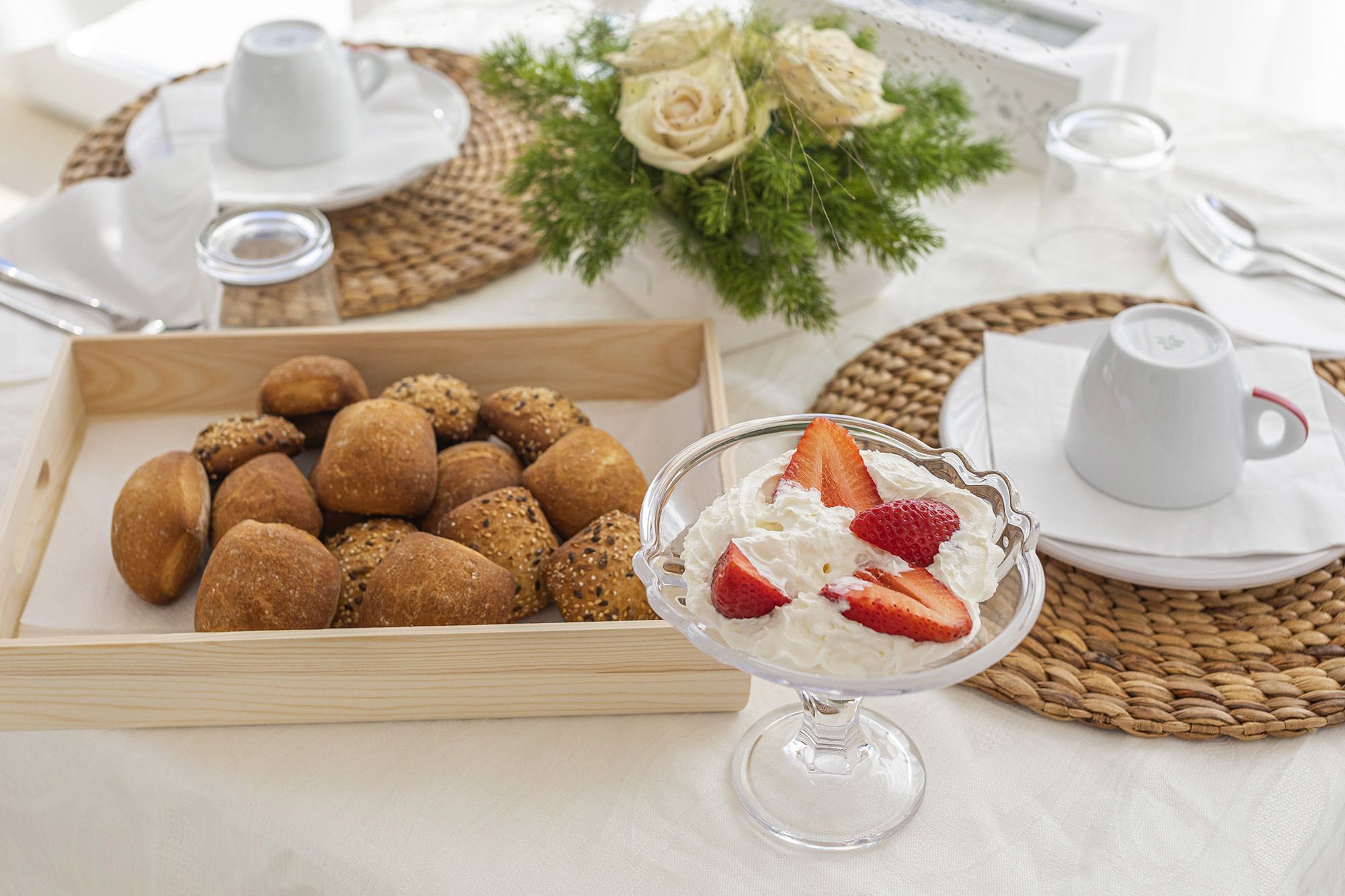 Une table mise pour le petit-déjeuner, avec un plateau de petits pains, un bol de dessert aux fraises et des tasses blanches.