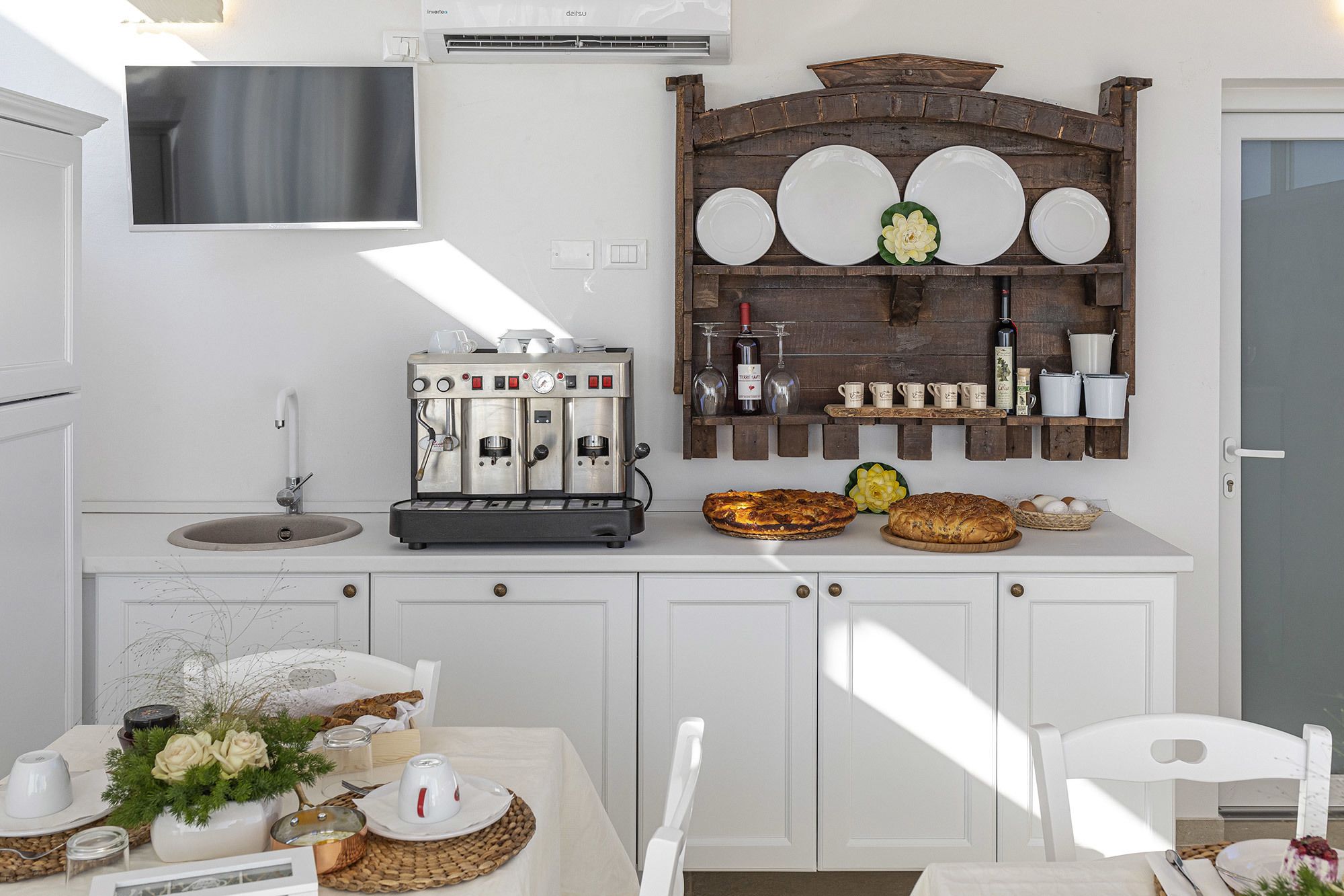 Una cocina moderna con armarios blancos, máquina de café espresso integrada, repisa de madera con platos y vasos, y un área de comedor con una mesa para dos.