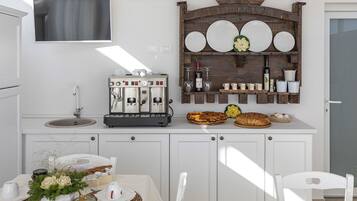 Una cocina moderna con armarios blancos, máquina de café espresso integrada, repisa de madera con platos y vasos, y un área de comedor con una mesa para dos.