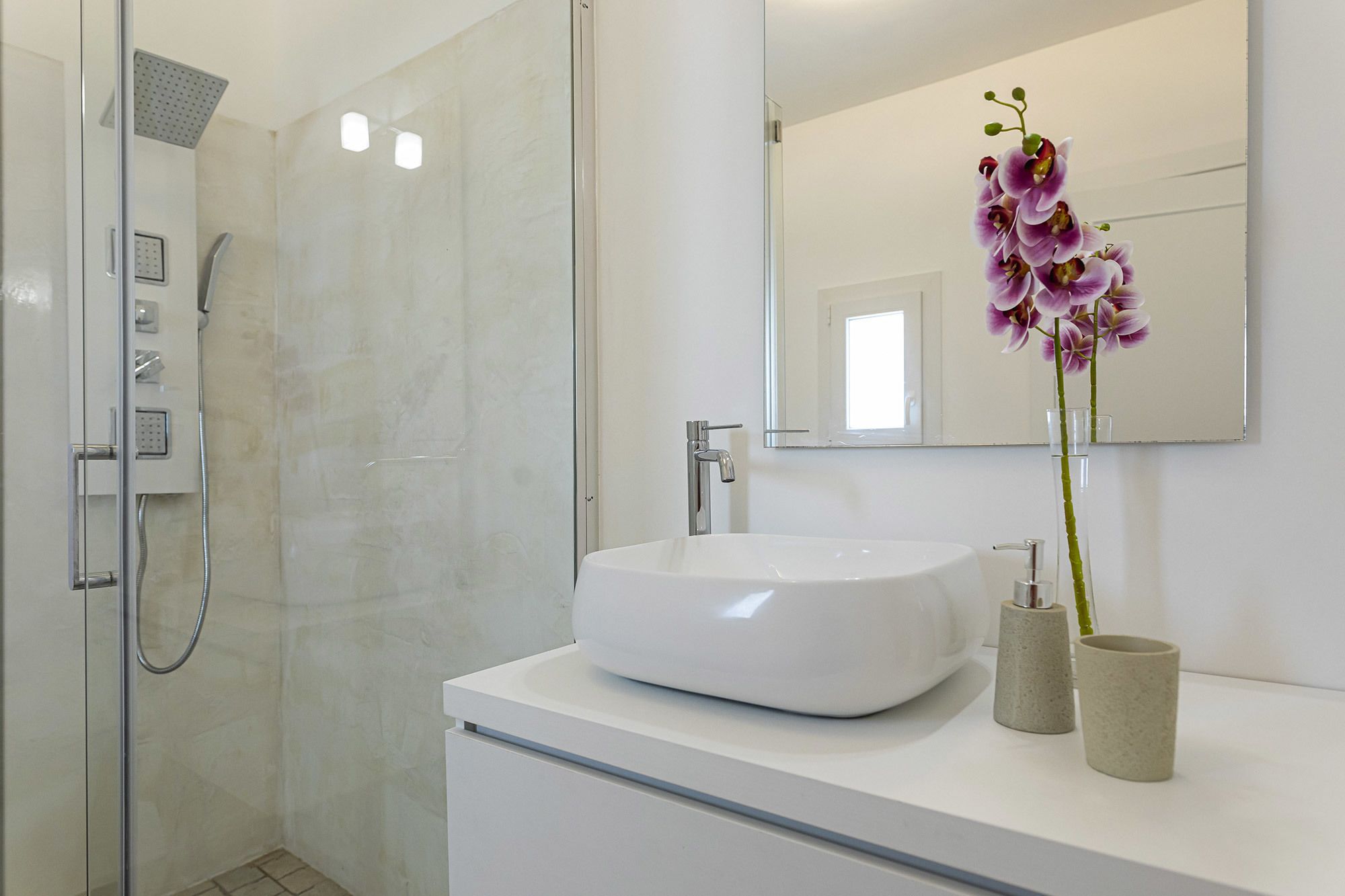 Un baño moderno con ducha envuelta en vidrio, un lavabo blanco y un jarrón con orquídeas moradas.