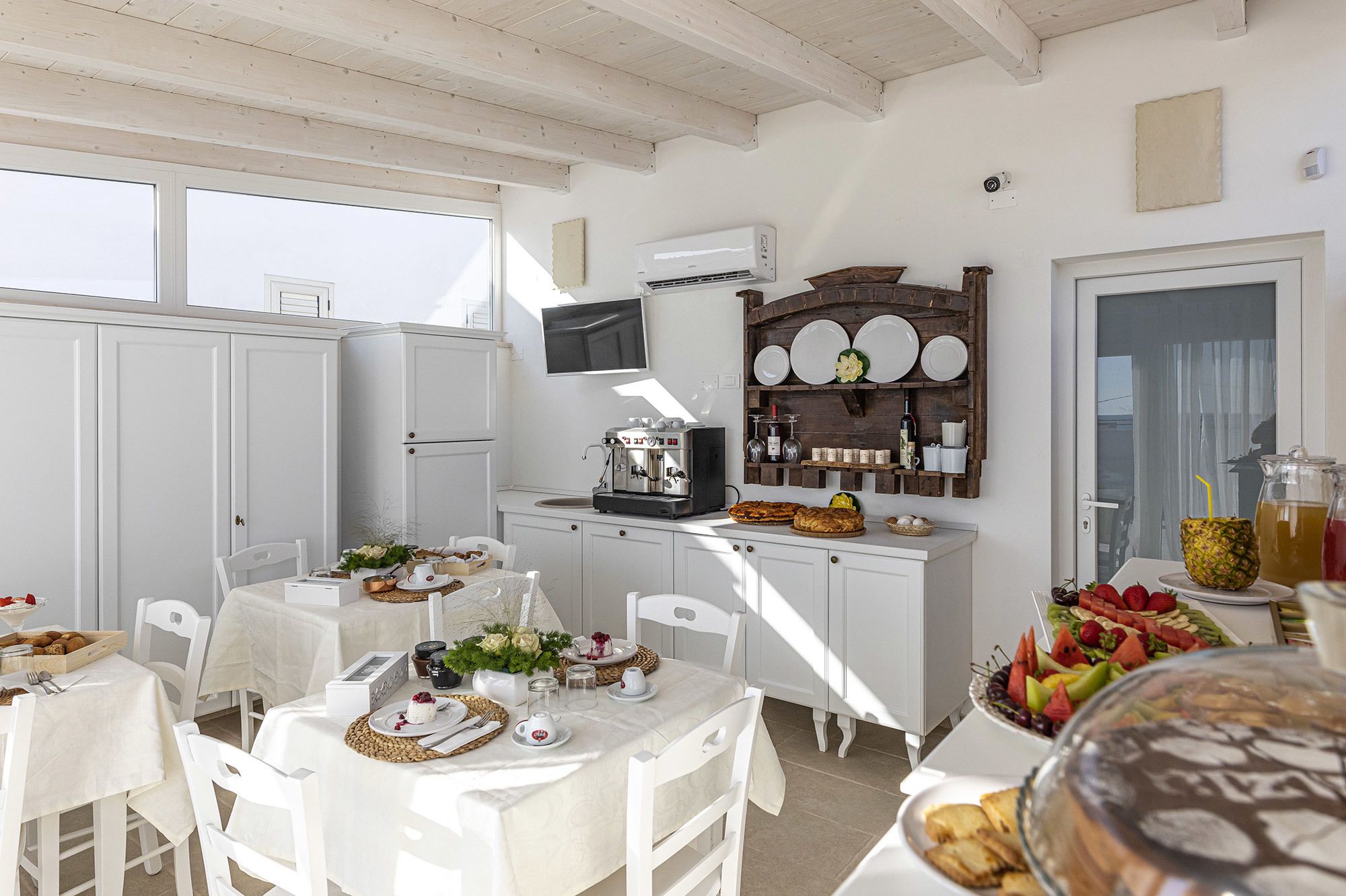 A bright, sunlit kitchen with white cabinetry, a coffee machine, and a fruit display.