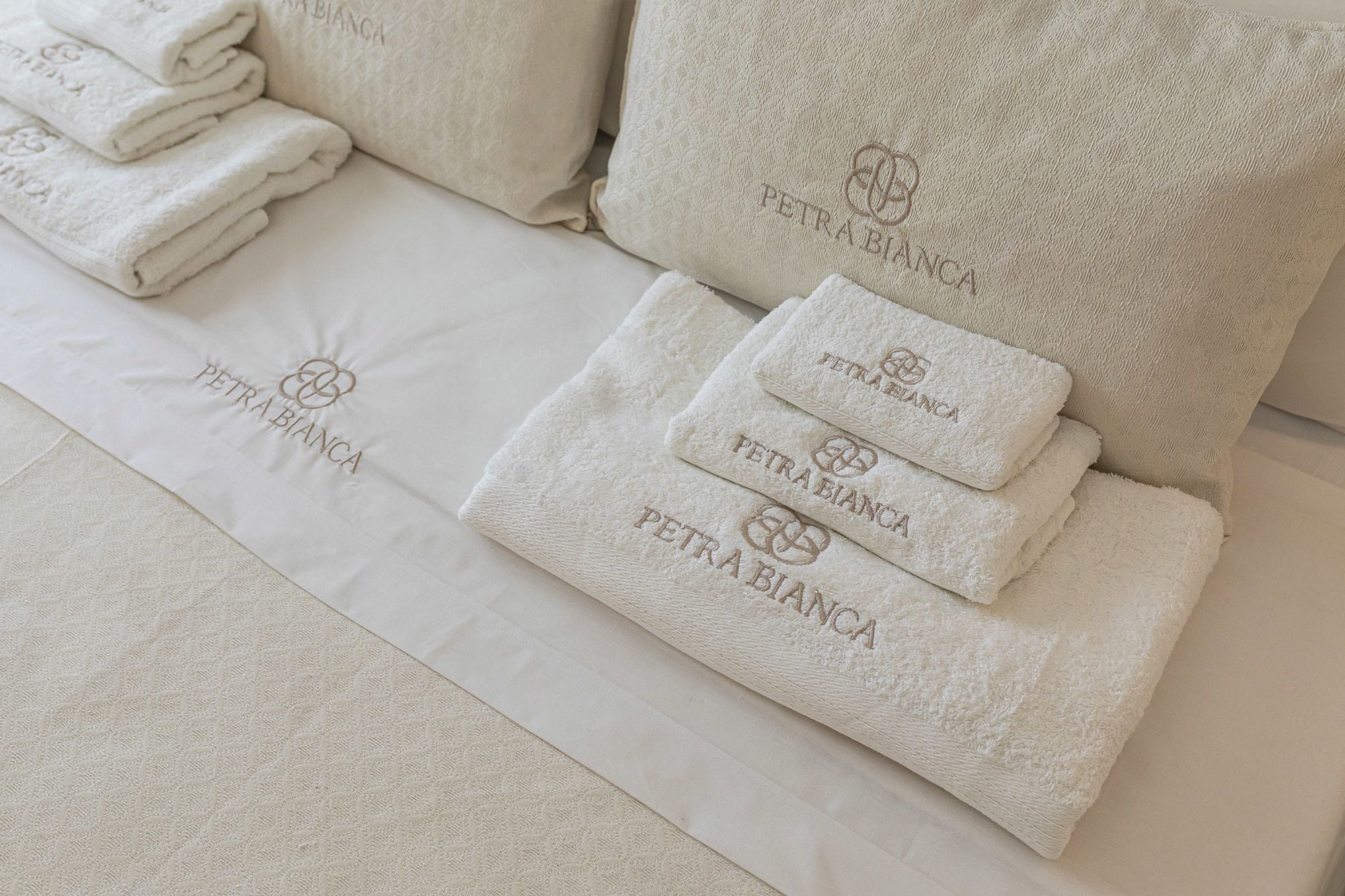 Una cama con ropa de cama y toallas blancas apiladas, cada una con el logo "PETRABIANCA".