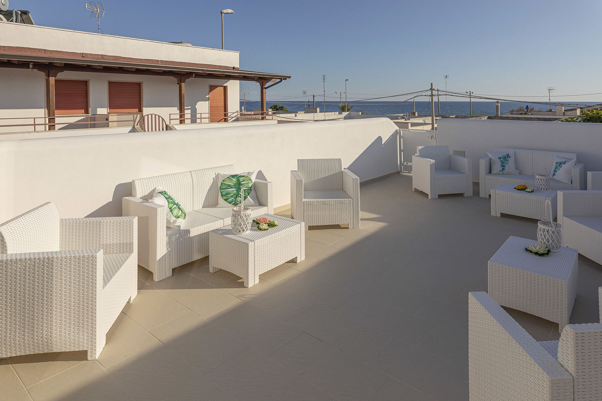 A rooftop terrace with white wicker furniture, including sofas and chairs, arranged on a tiled floor.