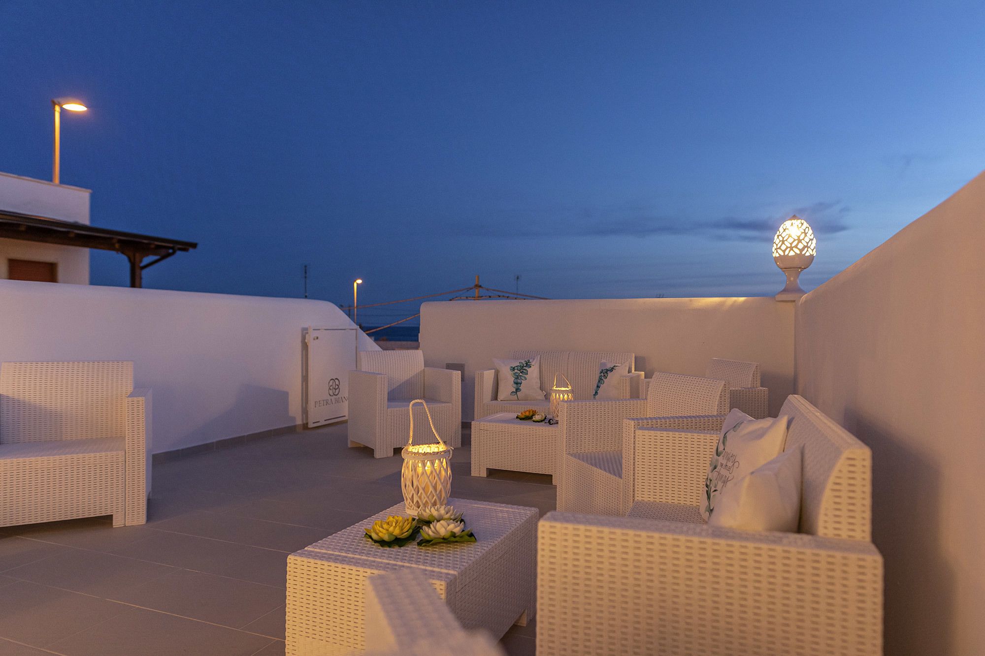 Terraza en la azotea con mobiliario de mimbre blanco, incluyendo sofás y sillones, dispuestos alrededor de mesas con adornos decorativos.