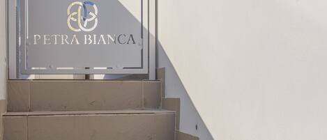Una señal con el texto “PETRA BIANCA” y un logo, colocada sobre una pared blanca con escalones que la suben.