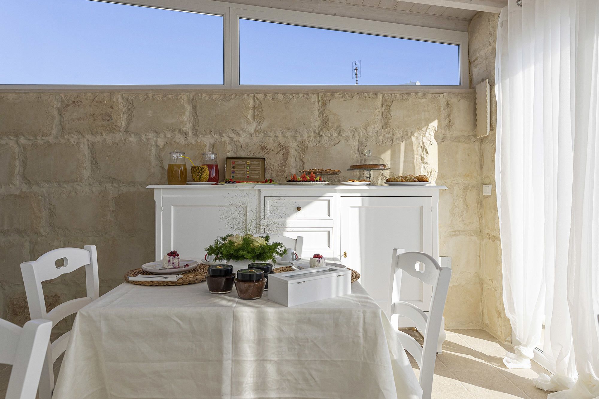 Un comedor con una mesa preparada para dos personas, un armario blanco con alimentos, y vista al cielo a través de una ventana.